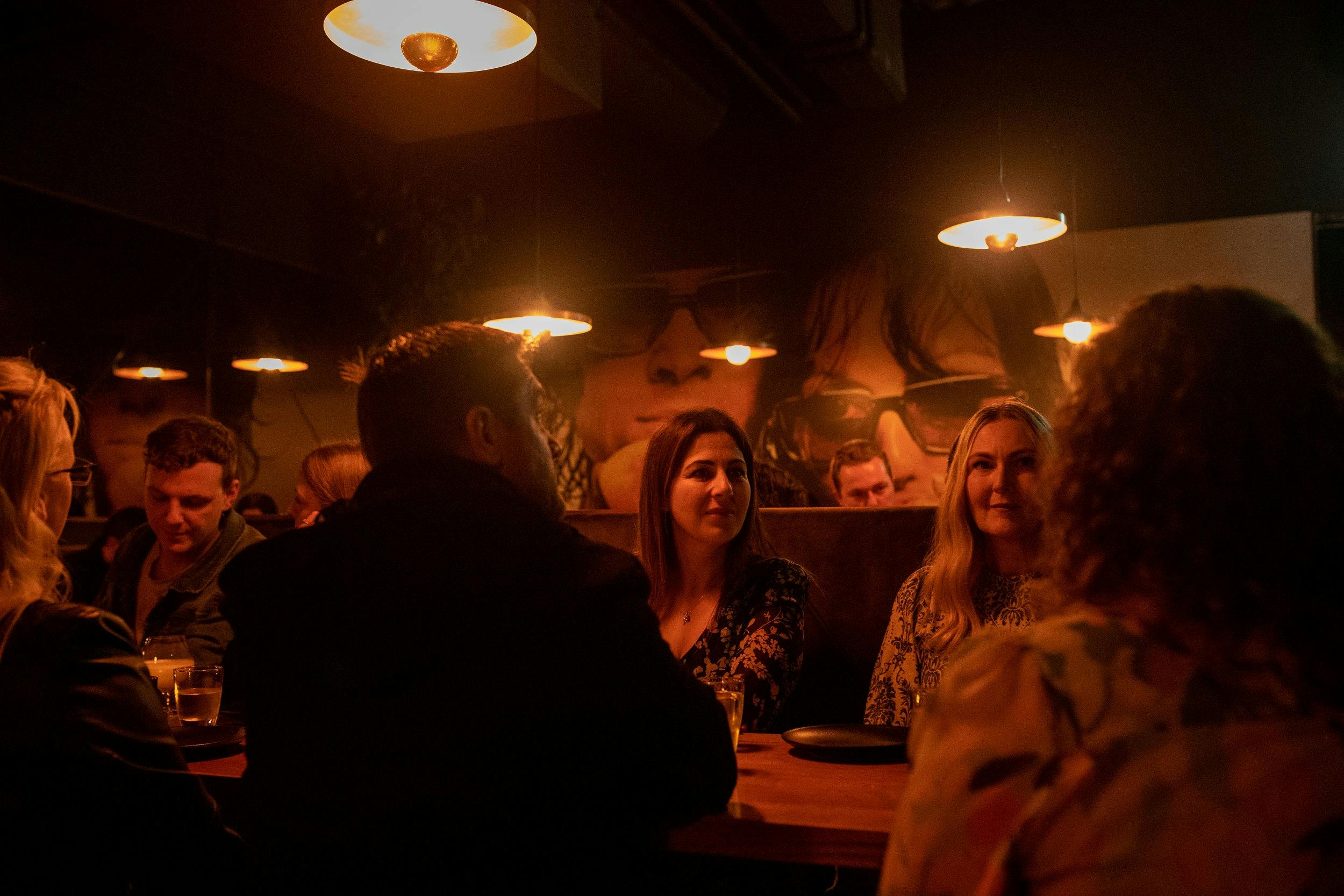A group of people sitting around a table.  The lighting is red.  Some people are facing away.