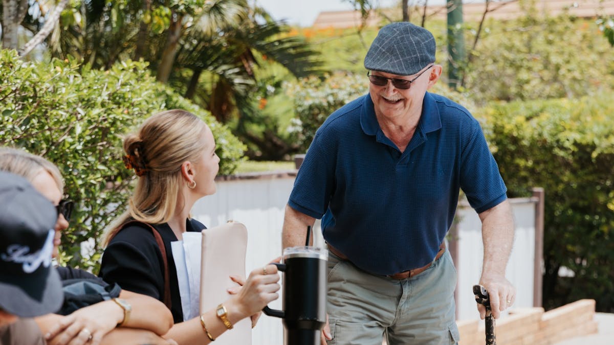 An older man with a cane smiling and chatting with a seated woman holding a drink, showing a warm, f