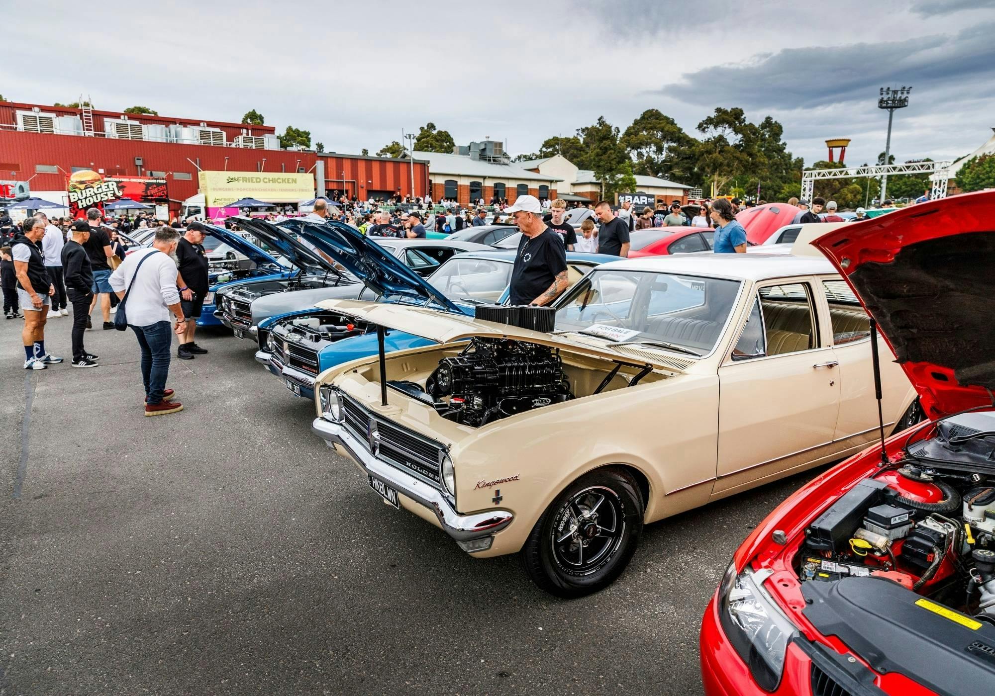Row of classic and modified cars with open bonnets, led by a cream supercharged Holden.