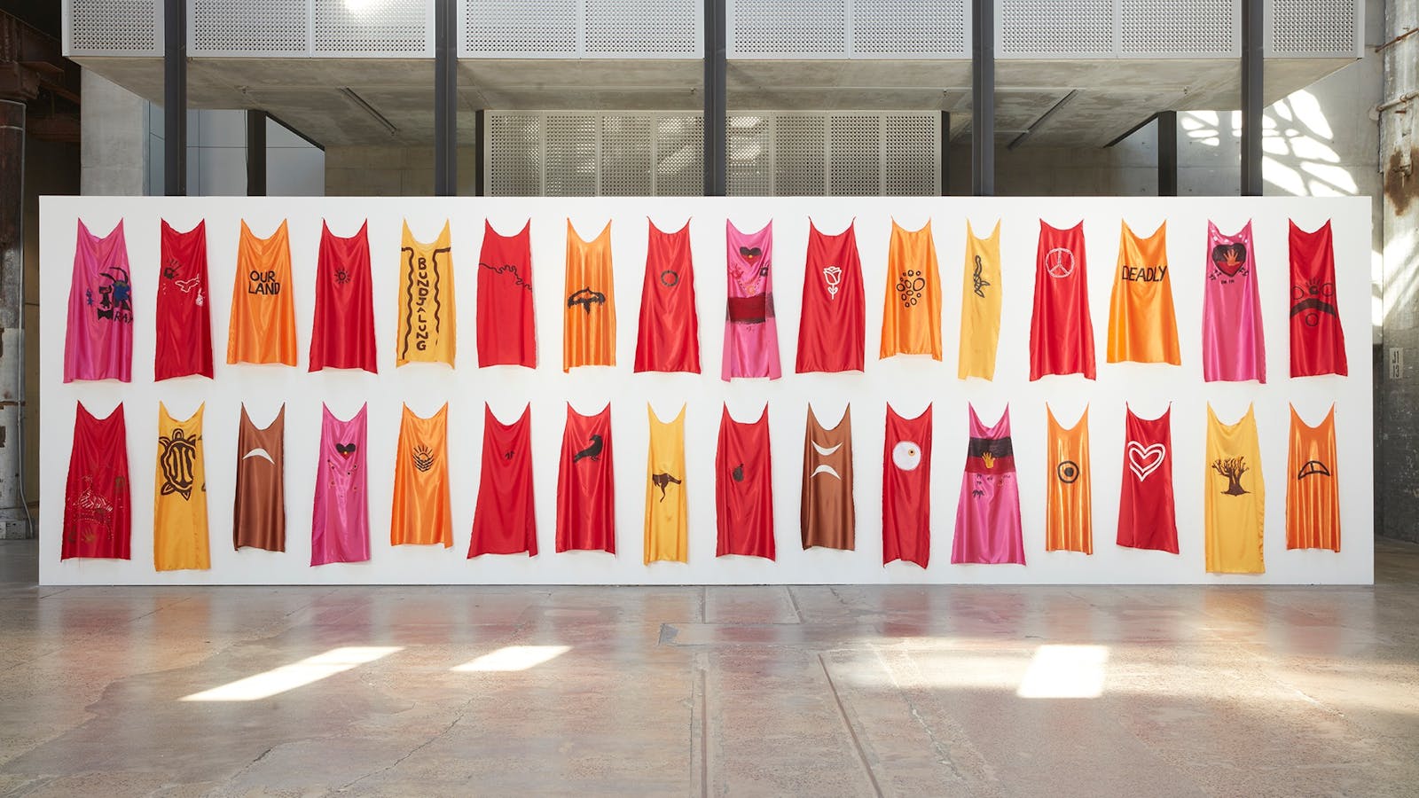 two rows of red, pink, orange, brown and yellow capes pinned to a white wall