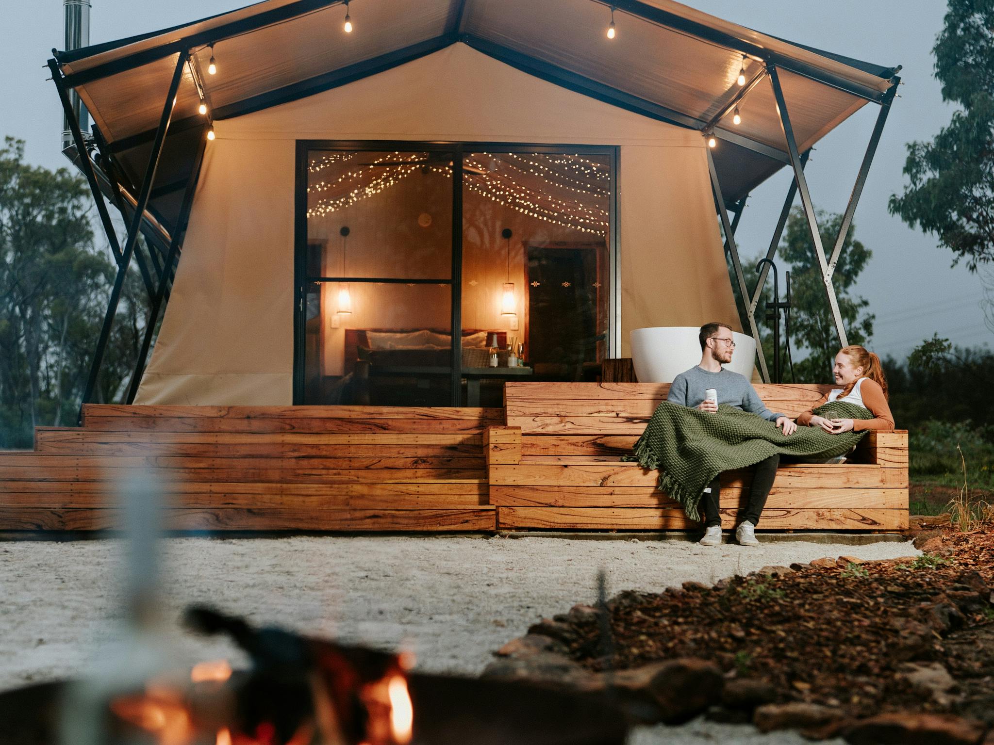 Couple relaxing on lounge at front of tent with fire pit in foreground