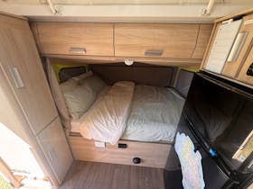 Queen bed made up with pale green bedding, overhead storage, cupboard to left and fridge to right