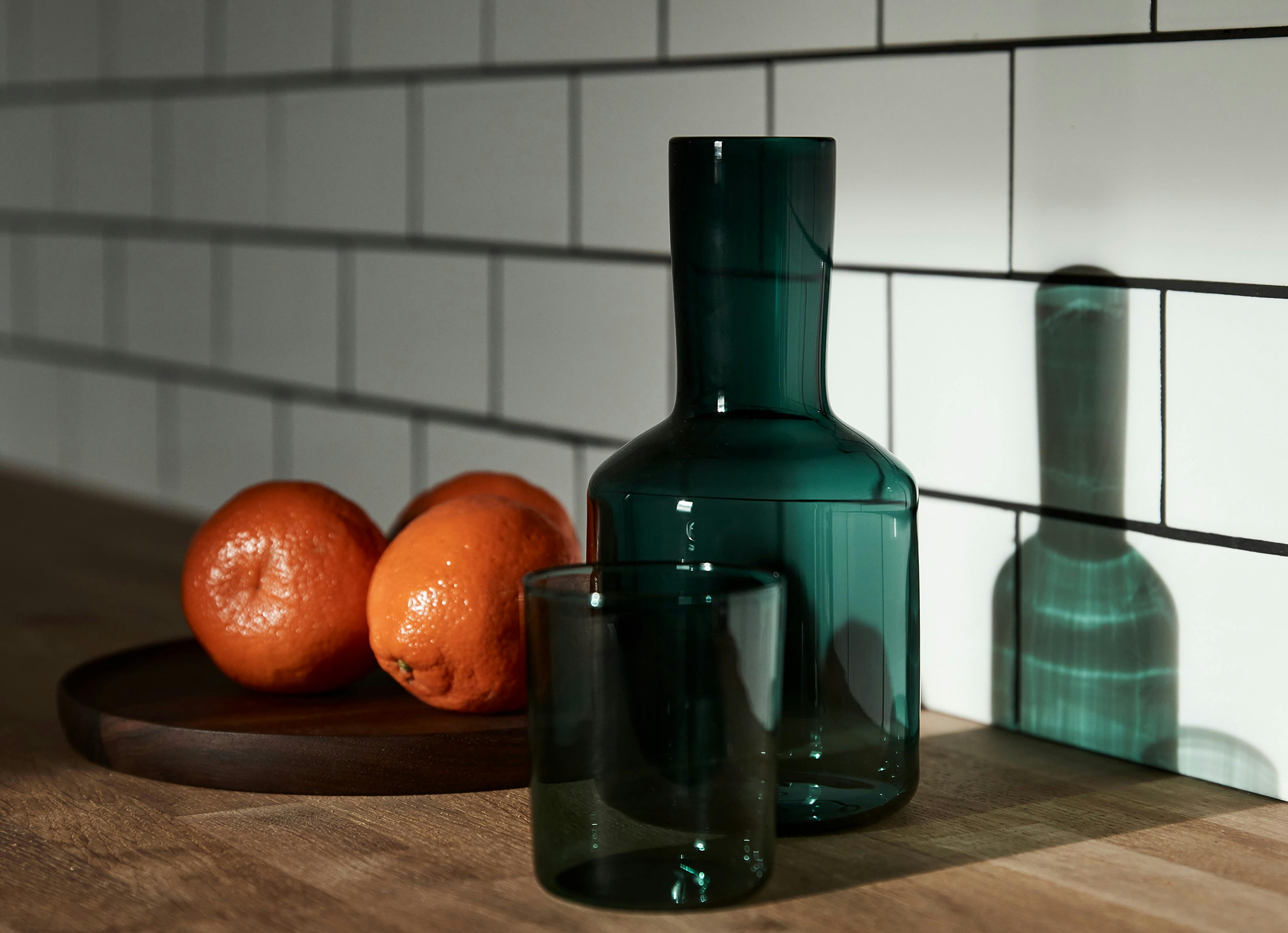 Close-up of green glassware and fresh oranges on a wooden surface with white subway tile backdrop