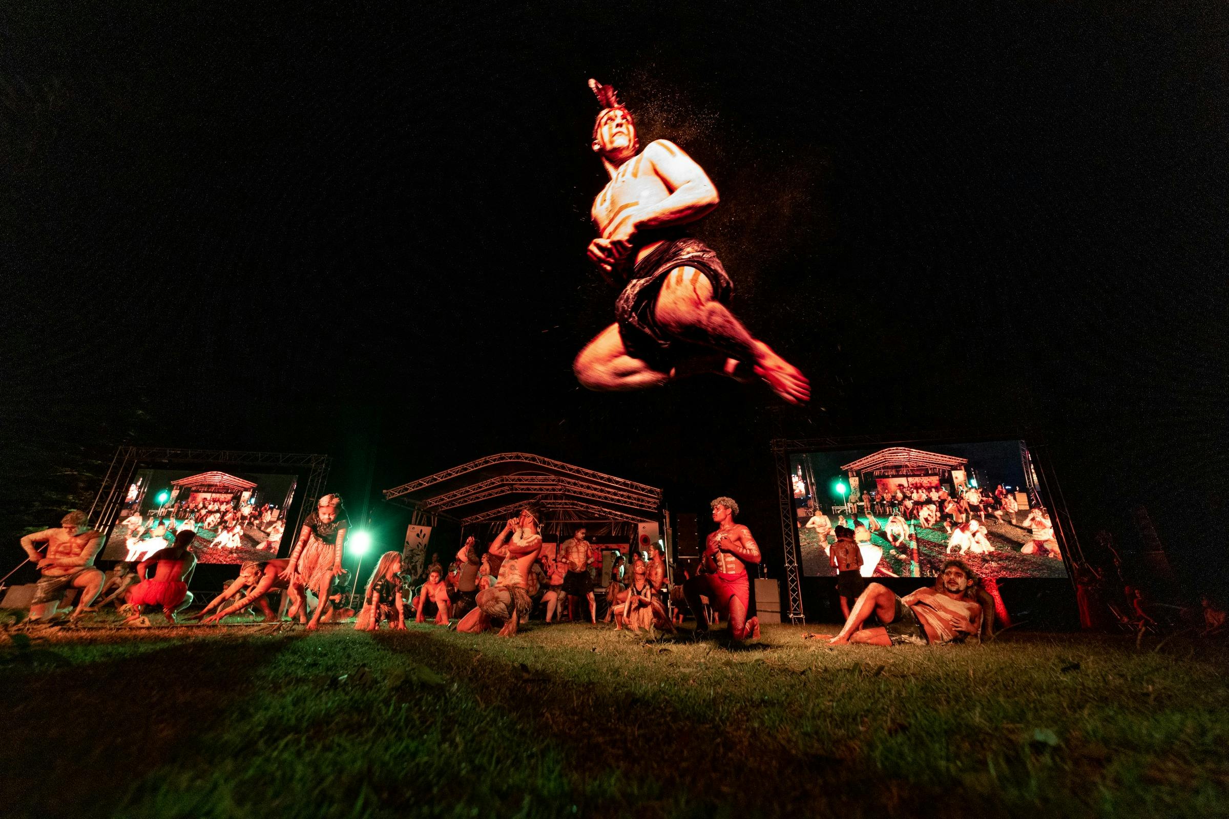 Warmly lit Aboriginal dancers perform on a grassy stage at night, the centre figure is airborne.