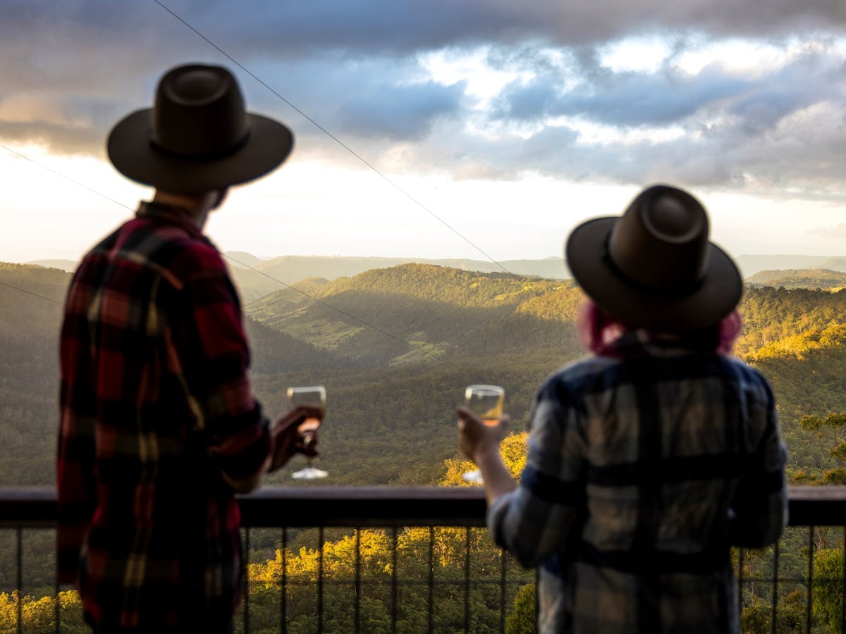A couple stand at a balcony railing with a glass of wine in their hand looking at the valley below
