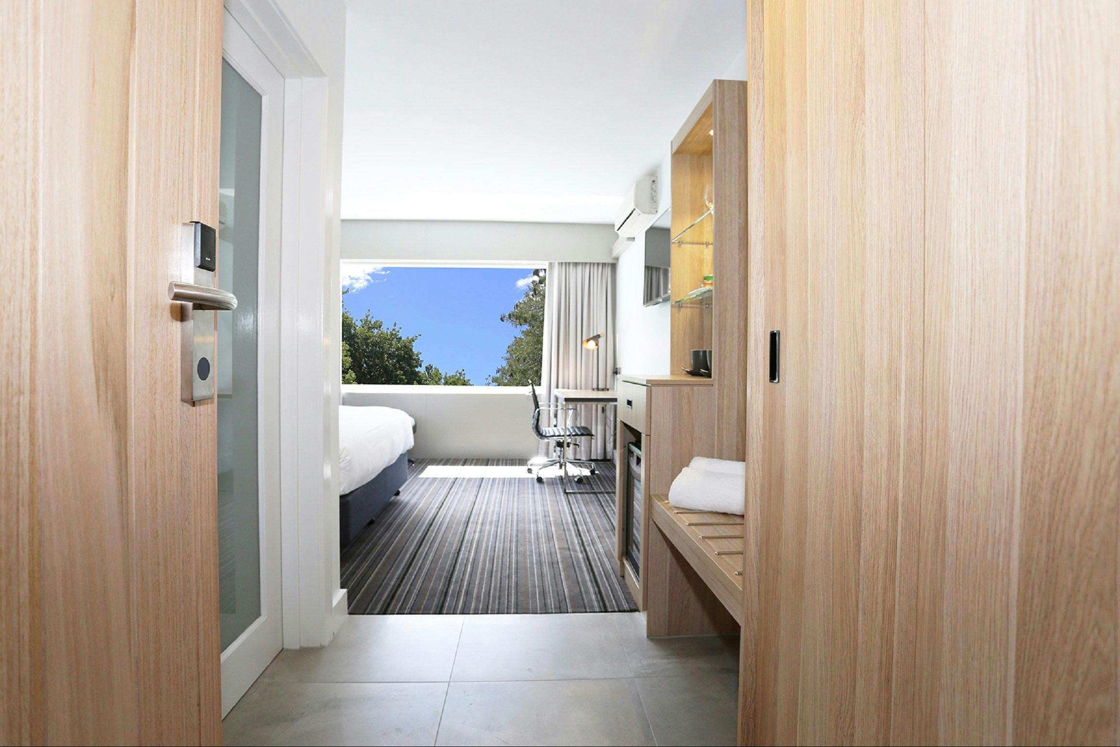 Entrance to hotel room, featuring desk, luggage rack and window with sky view.