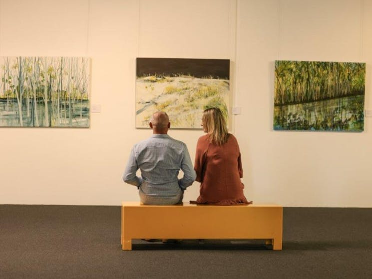 A bald man and blonde woman sit on a mustard bench looking at 3 artworks on a white wall.