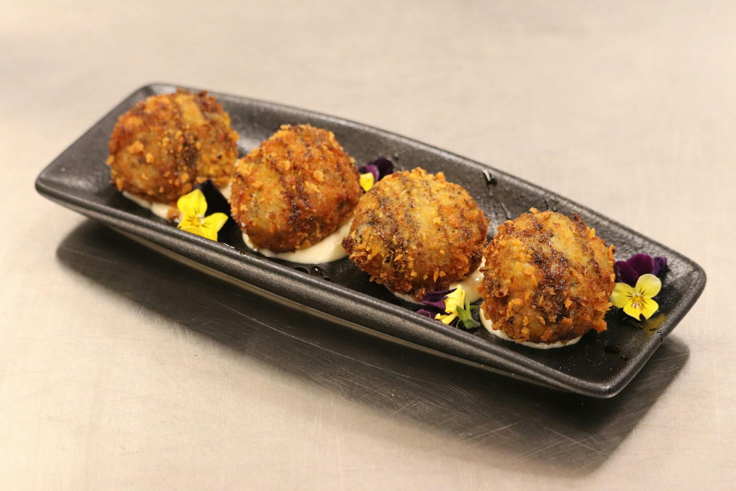 Golden fried croquettes served on a black plate with sauce and edible flowers.