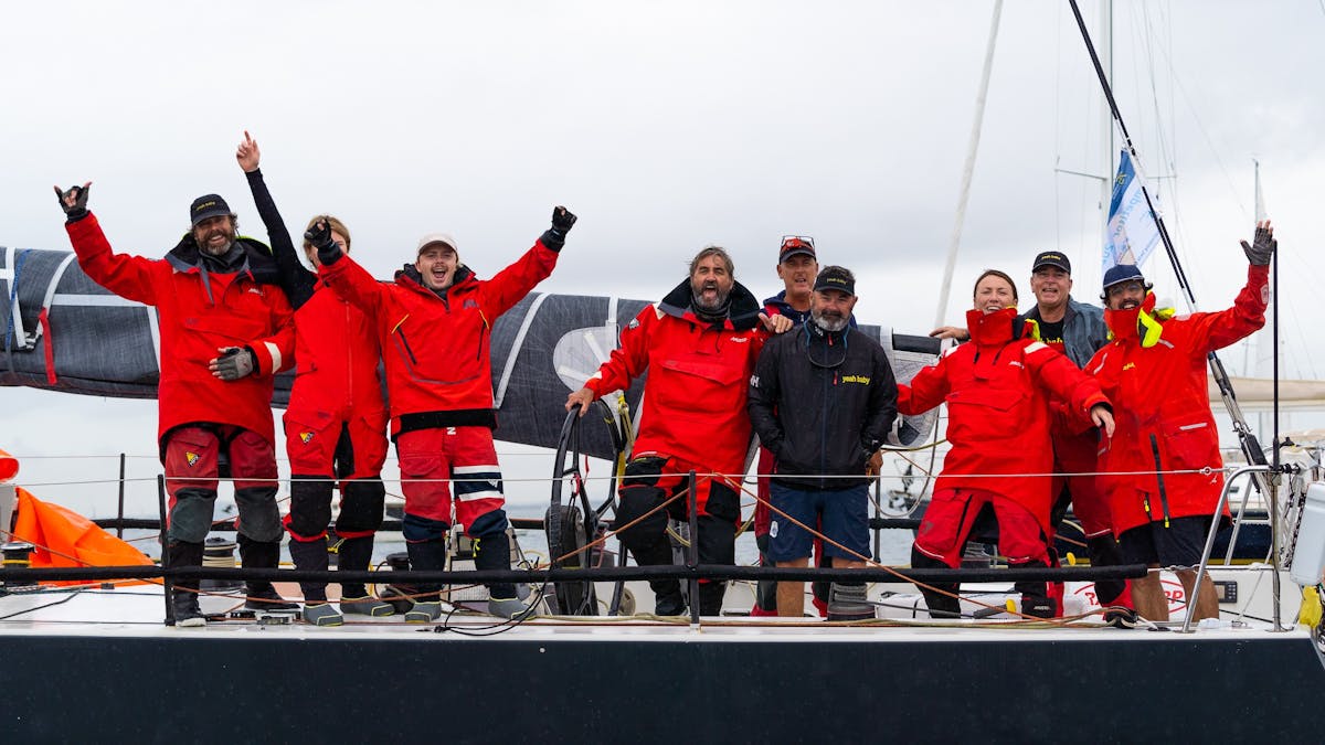 Sailors standing on the side of their yacht in red jackets waving at the camera at race finish