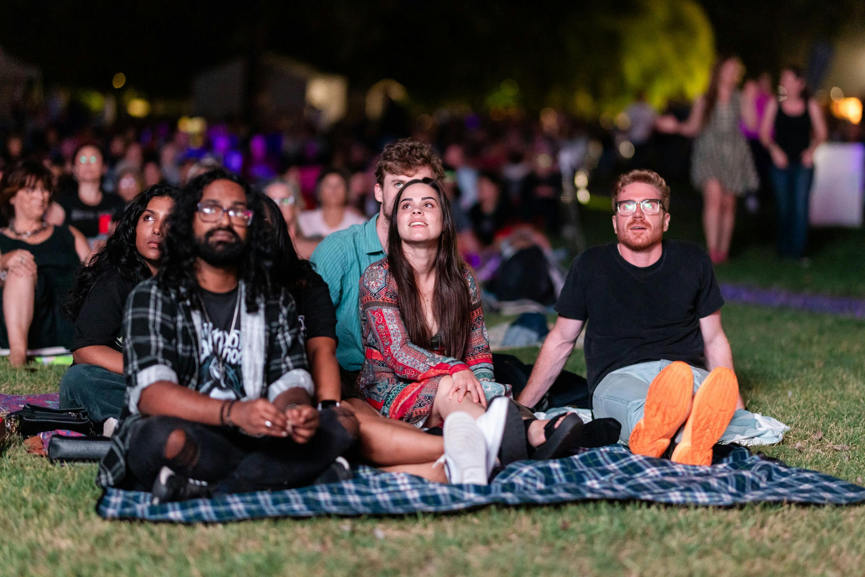 People sitting on grass, looking up at stage as they watch outdoor performance