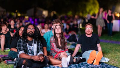 People sitting on grass, looking up at stage as they watch outdoor performance