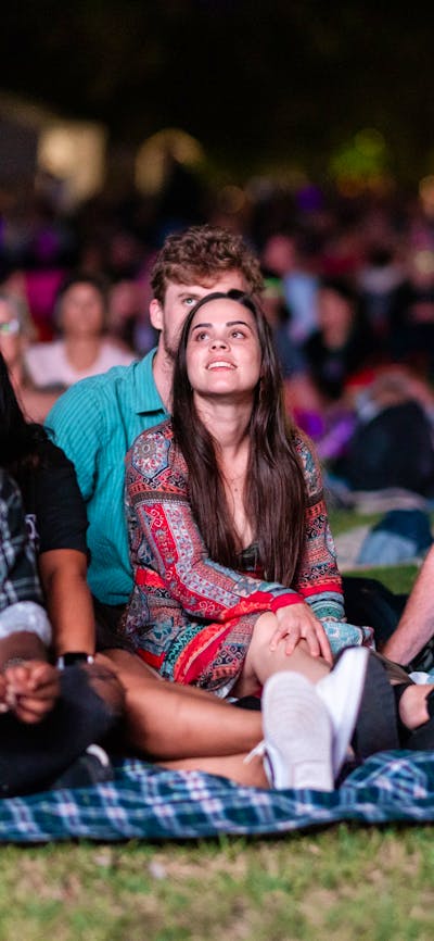 People sitting on grass, looking up at stage as they watch outdoor performance