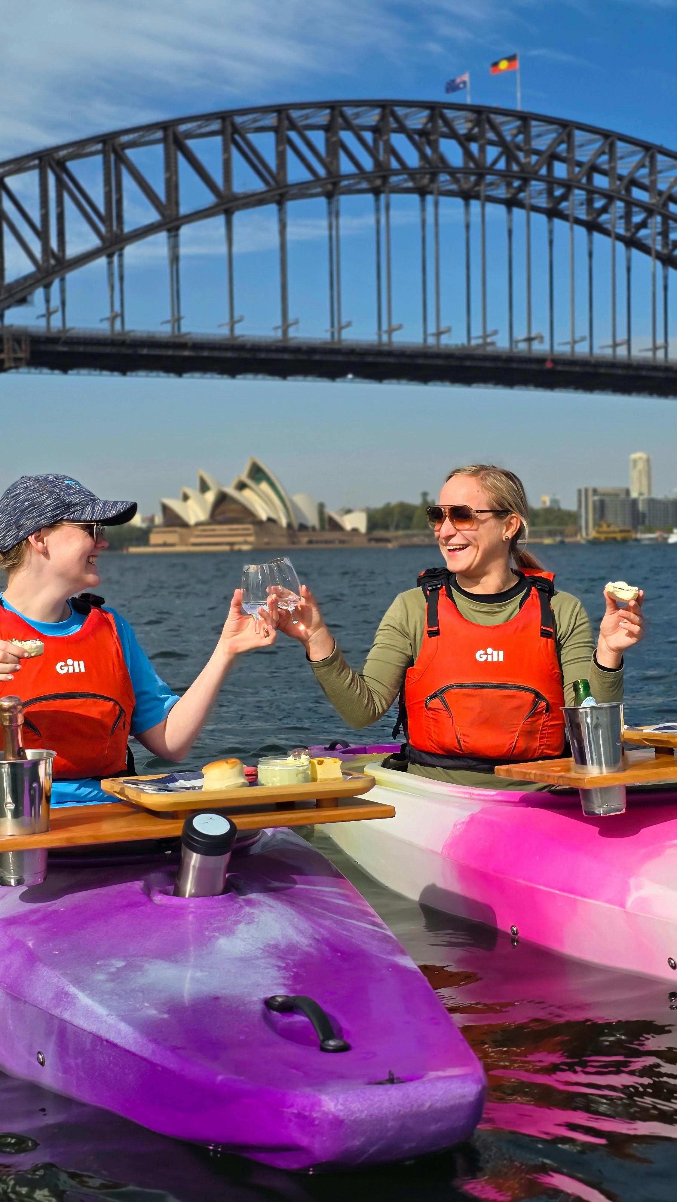 Two ladies clinking glasses with scones in hand in their kayaks with Sydney Opera House behind them.
