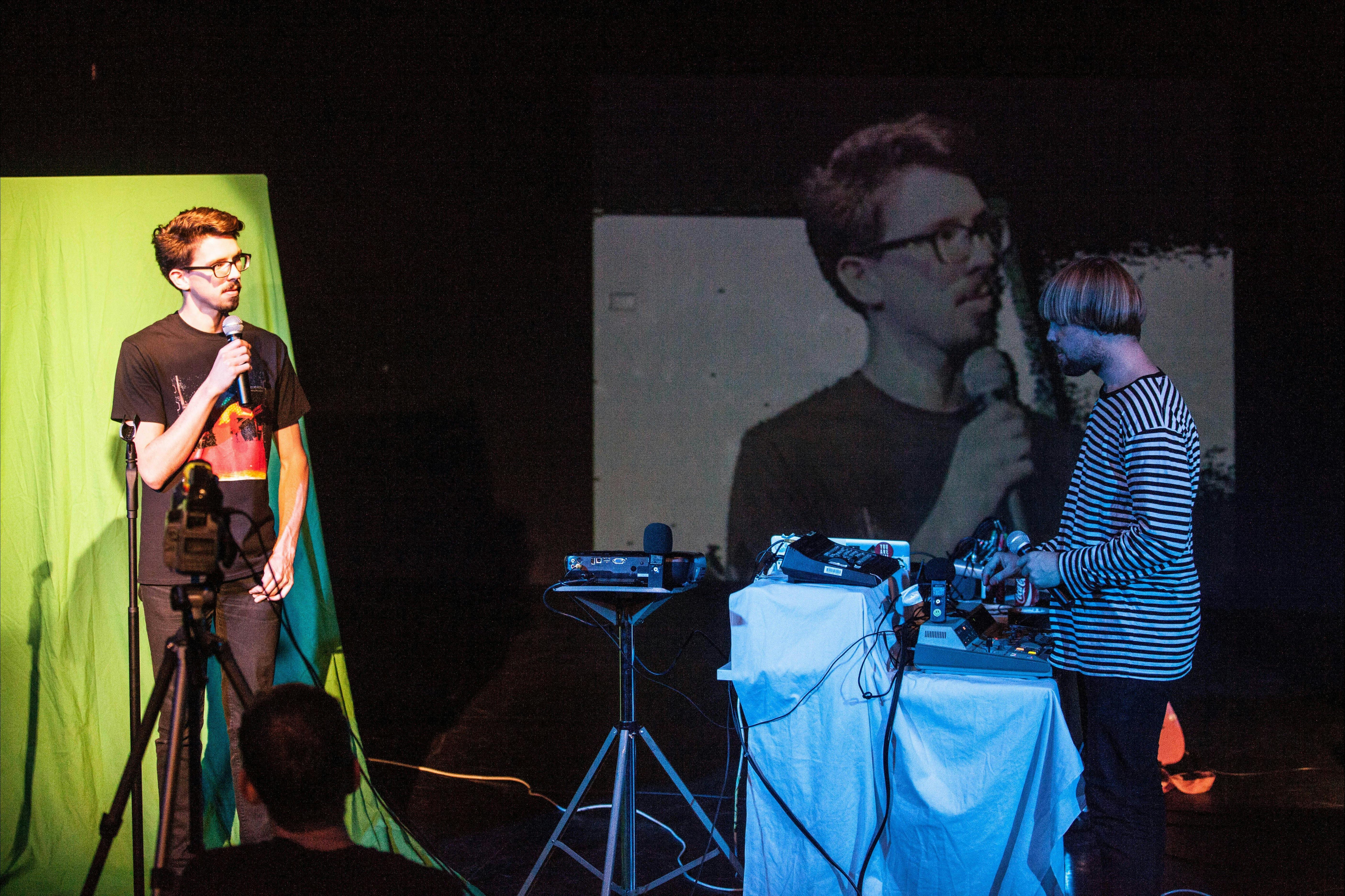 Two people in a blackbox theatre - man standing in front of green screen speaking into a microphone.