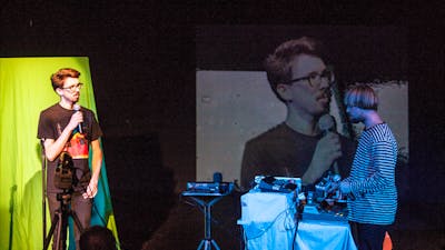 Two people in a blackbox theatre - man standing in front of green screen speaking into a microphone.