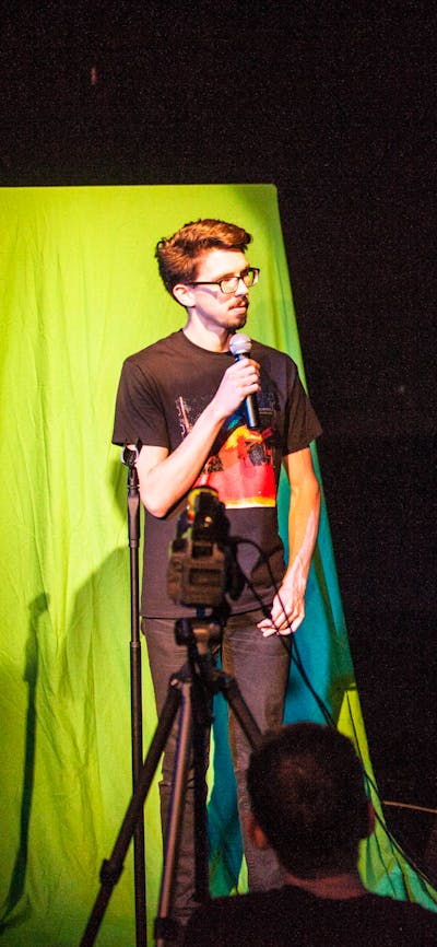 Two people in a blackbox theatre - man standing in front of green screen speaking into a microphone.