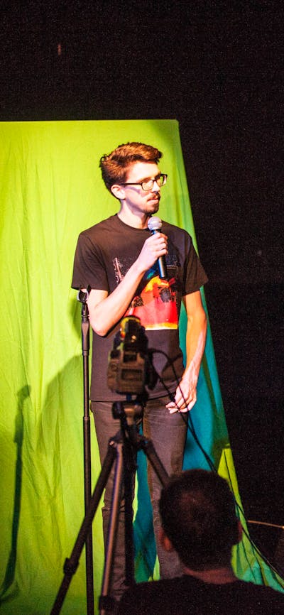 Two people in a blackbox theatre - man standing in front of green screen speaking into a microphone.