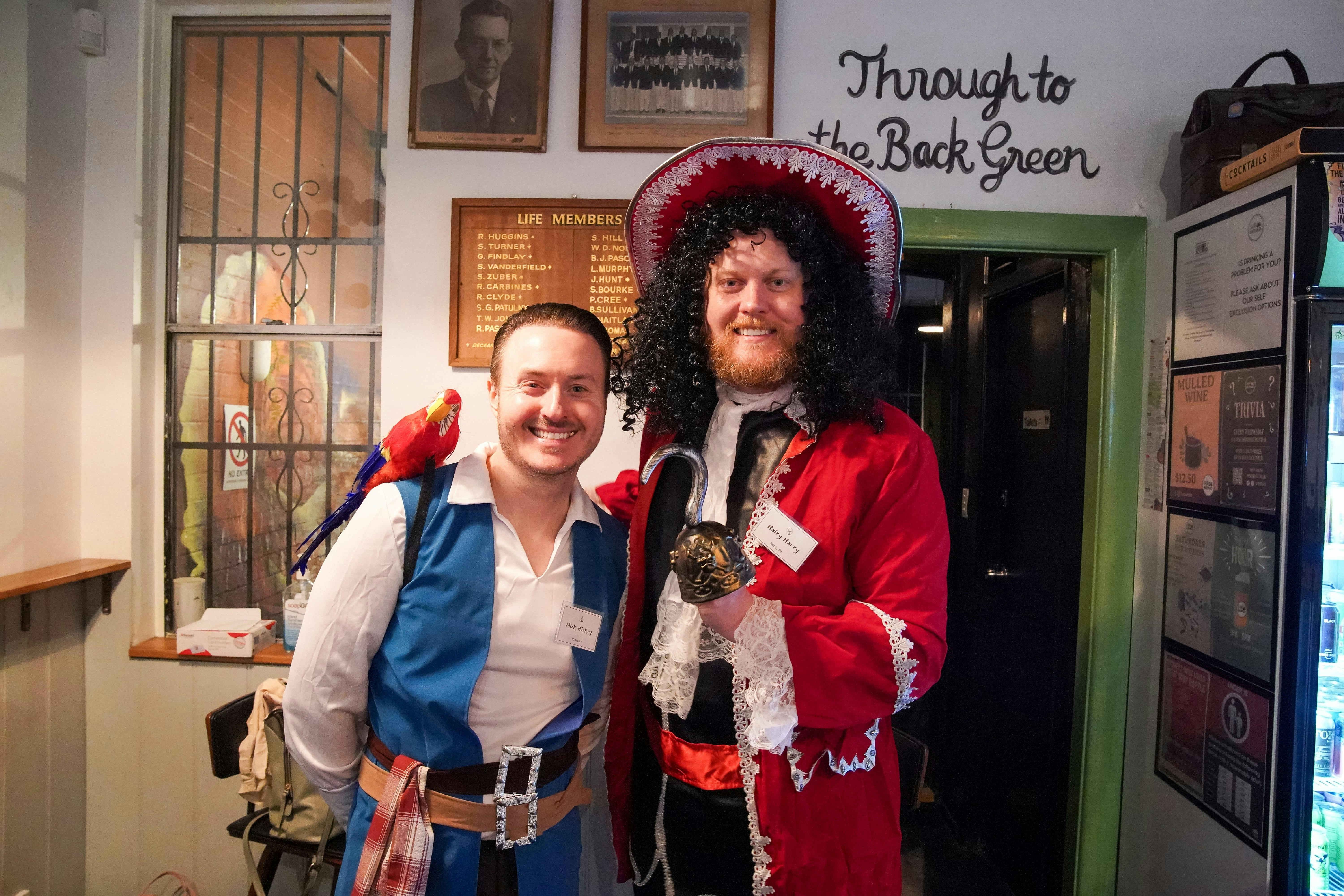 Two men in pirate outfits, one in blue, one in red, smile and pose for the camera