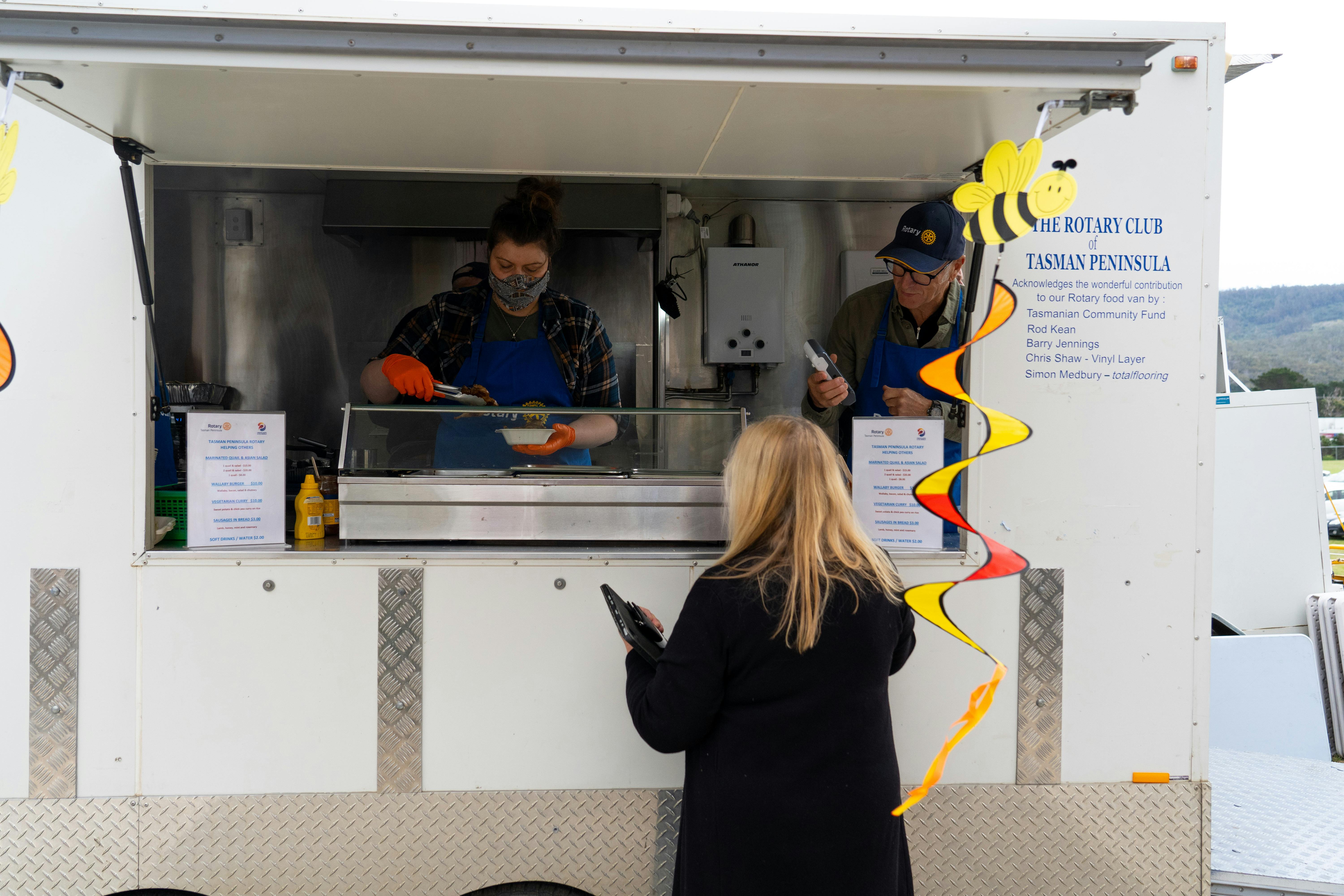 Two Rotarians inside the Rotary van serving up yummy food to a lady enjoying the fine food on offer