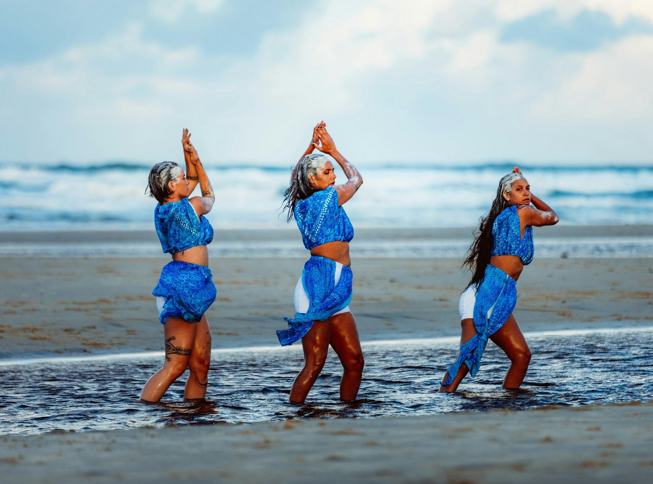 Three women in blue outfits dance in shallow seawater on a beach, with ocean waves behind them.