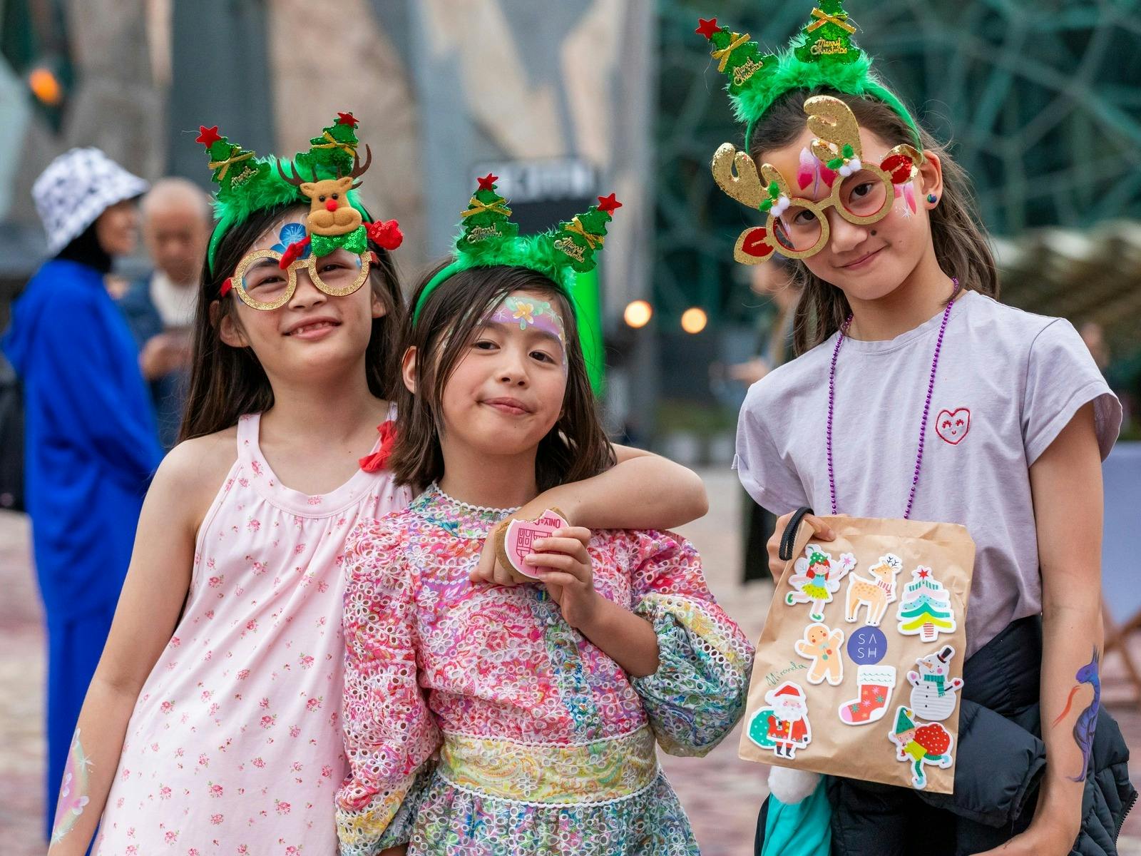 Three children pose for a photo wearing Christmas tree headbands at Fed Square