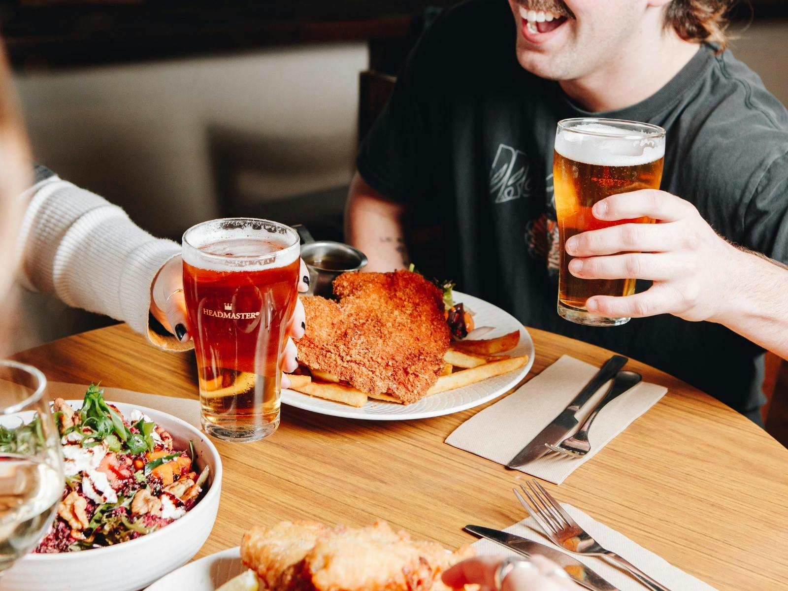 Two people holding glasses of beer over a table with a chicken schnitzel, chips and salad at Great S
