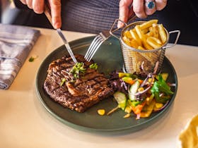 A plate of grilled steak, basket of chips and side salad, with a knife and fork poised to tuck in