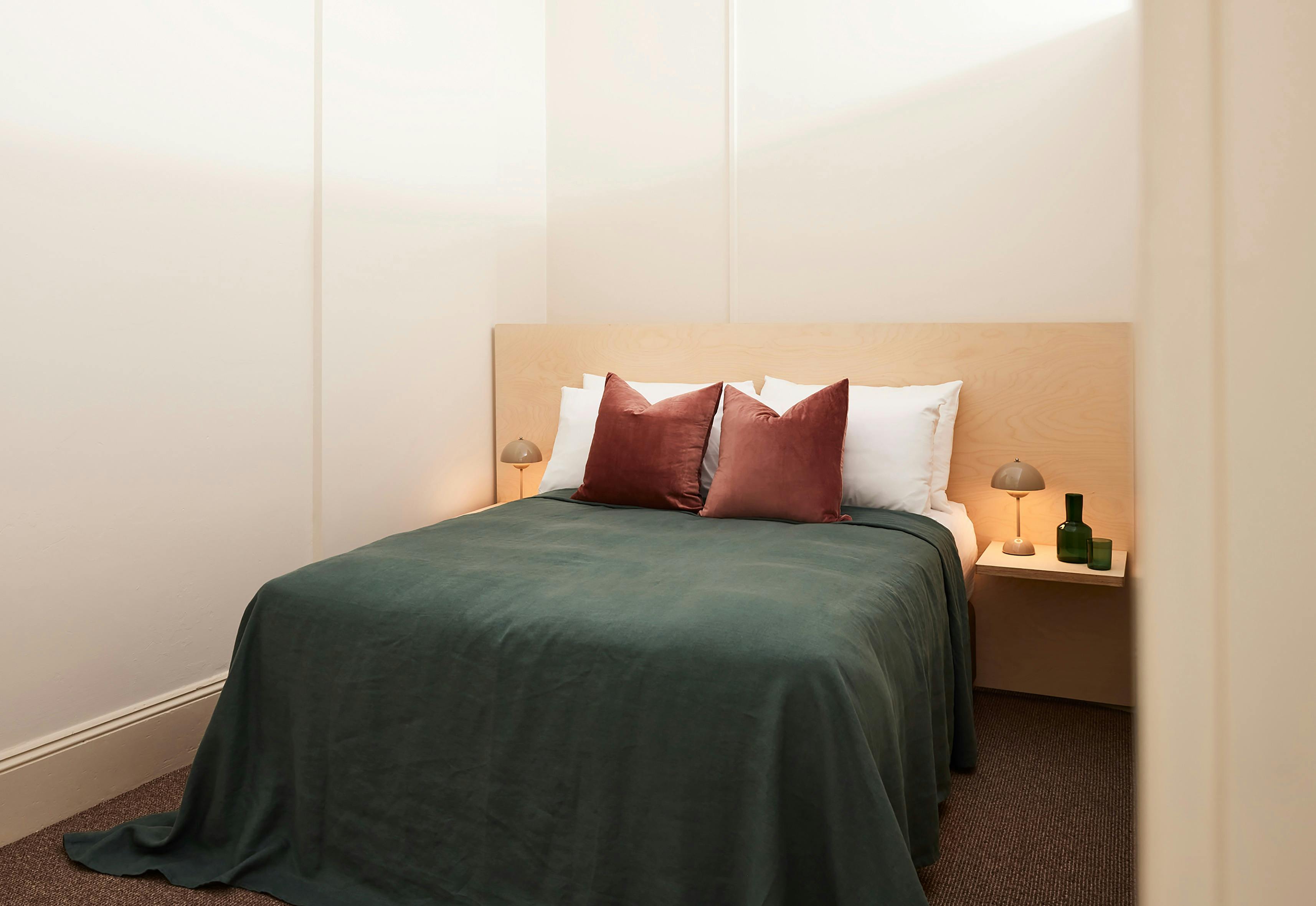 Minimalist bedroom with light timber headboard, green bedspread, and rust-colored accent pillows