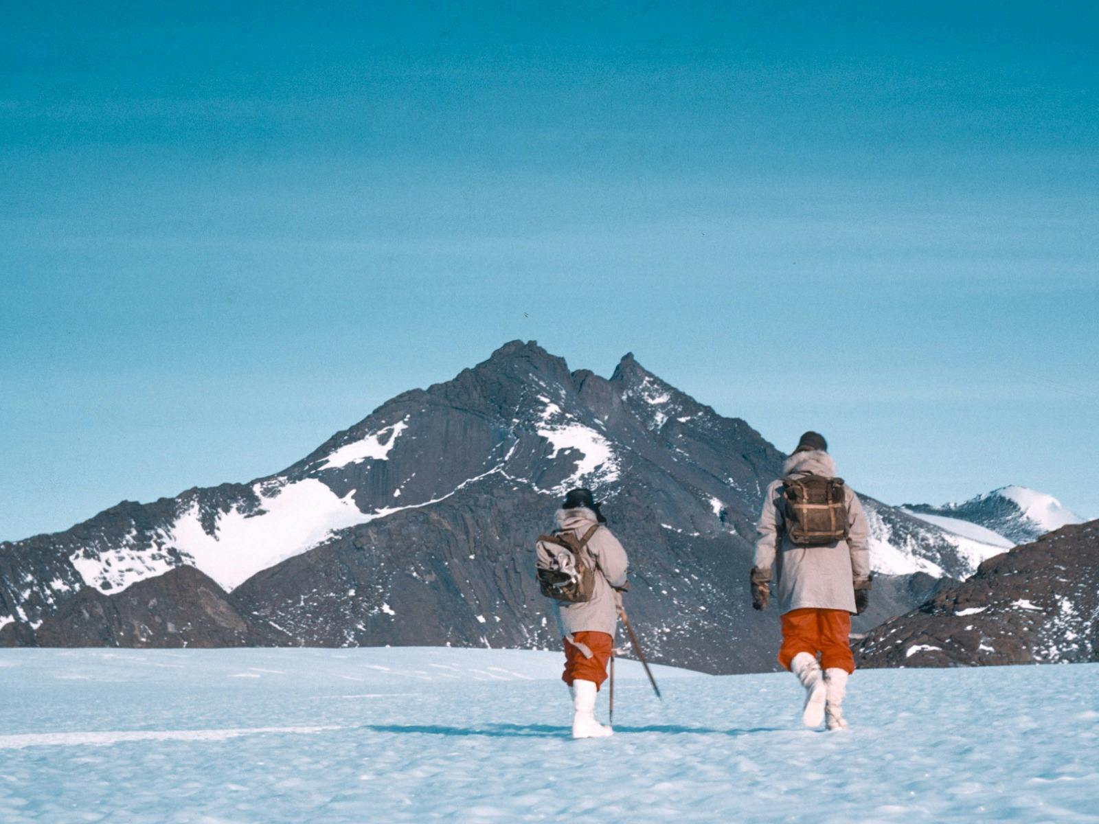 Two hikers trekking across a snow-covered landscape with a mountain in the background.