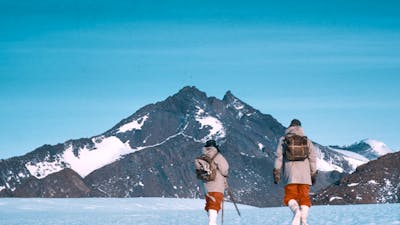 Two hikers trekking across a snow-covered landscape with a mountain in the background.