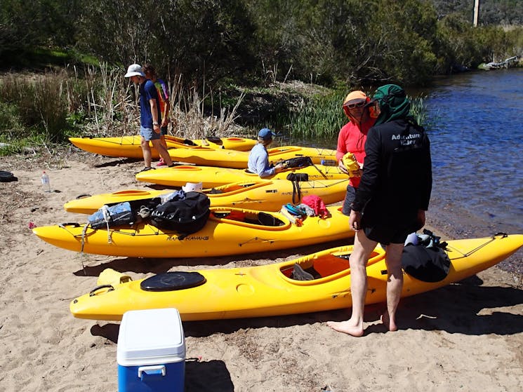 six yellow kayaks are on the bank of the snowy river with four people loading gear into them