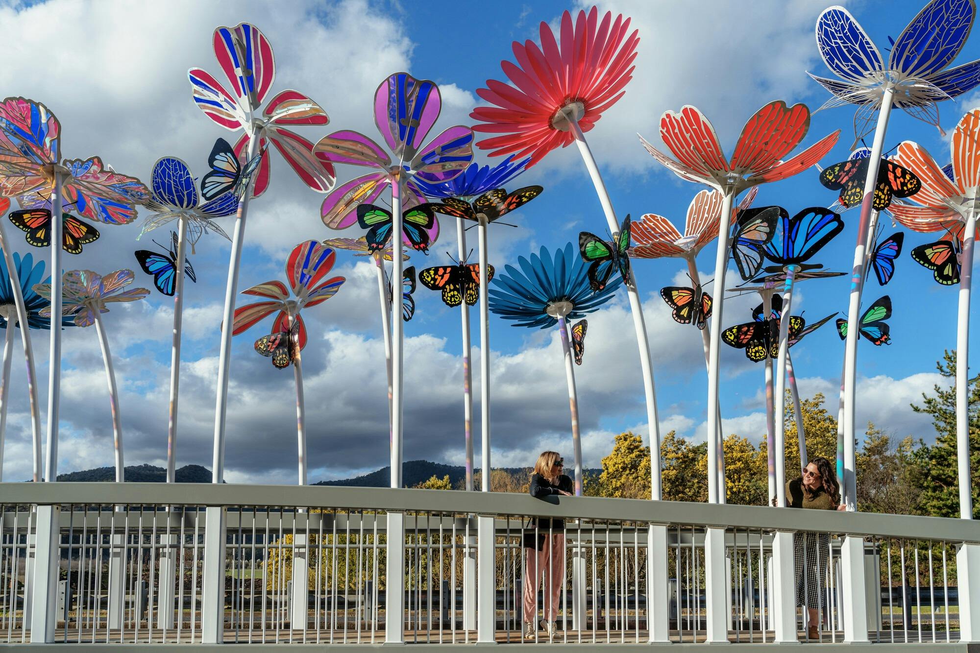 Two women leaning on a white bridge with tall colourful flower sculptures along its edges
