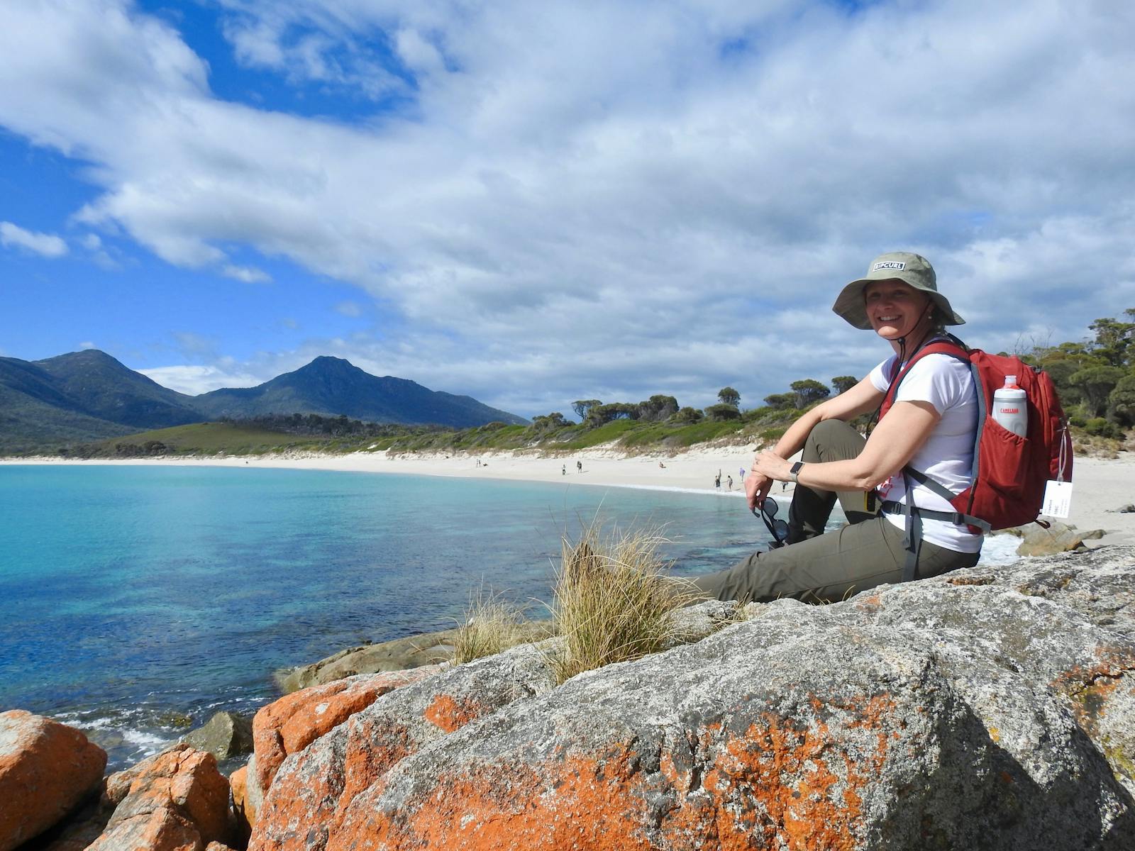 A smiling woman sits on lichen-covered rocks overlooking a turquoise bay