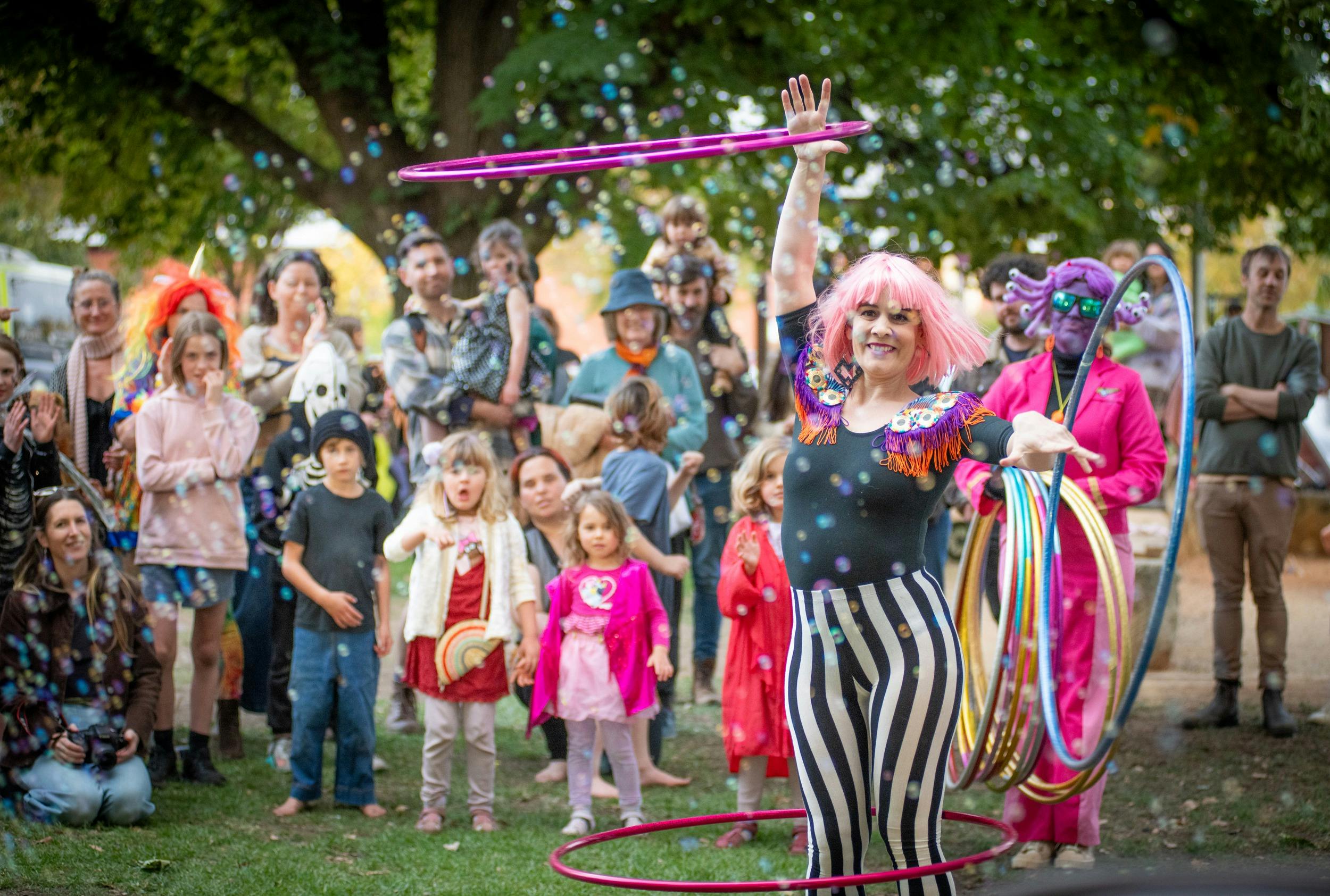 Hula Hooper in a pink wig performing in a park in front of an audience with bubbles in the air
