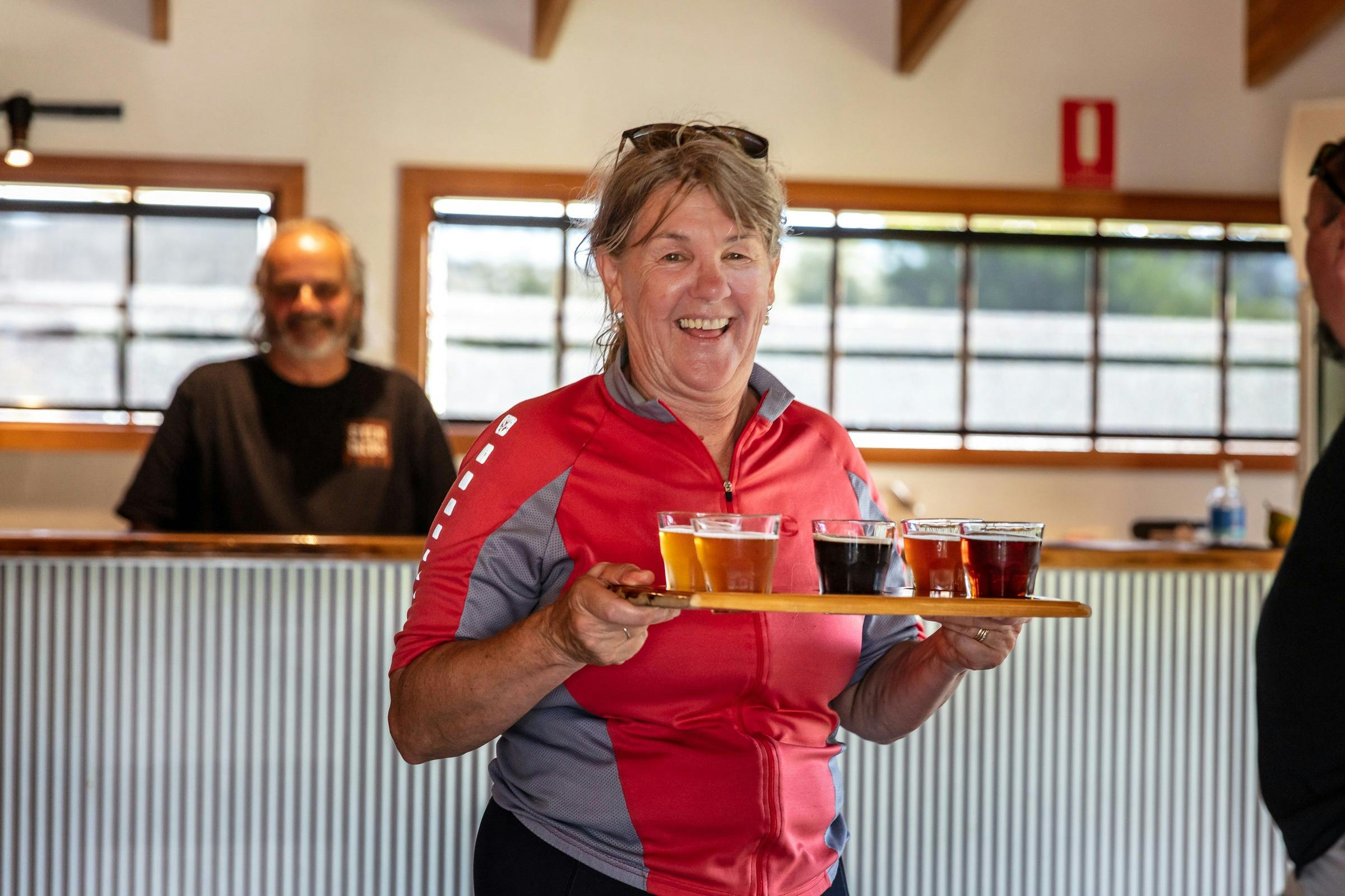 A woman in sportswear walking away from a bar with five glasses of different beers on a wooden tray.