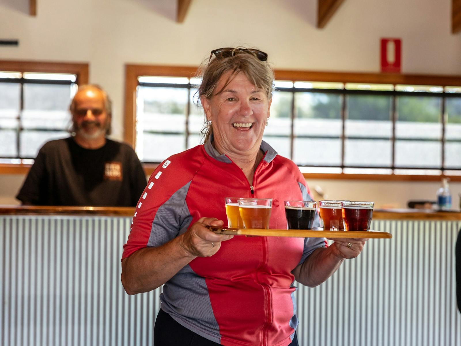 A woman in sportswear walking away from a bar with five glasses of different beers on a wooden tray.