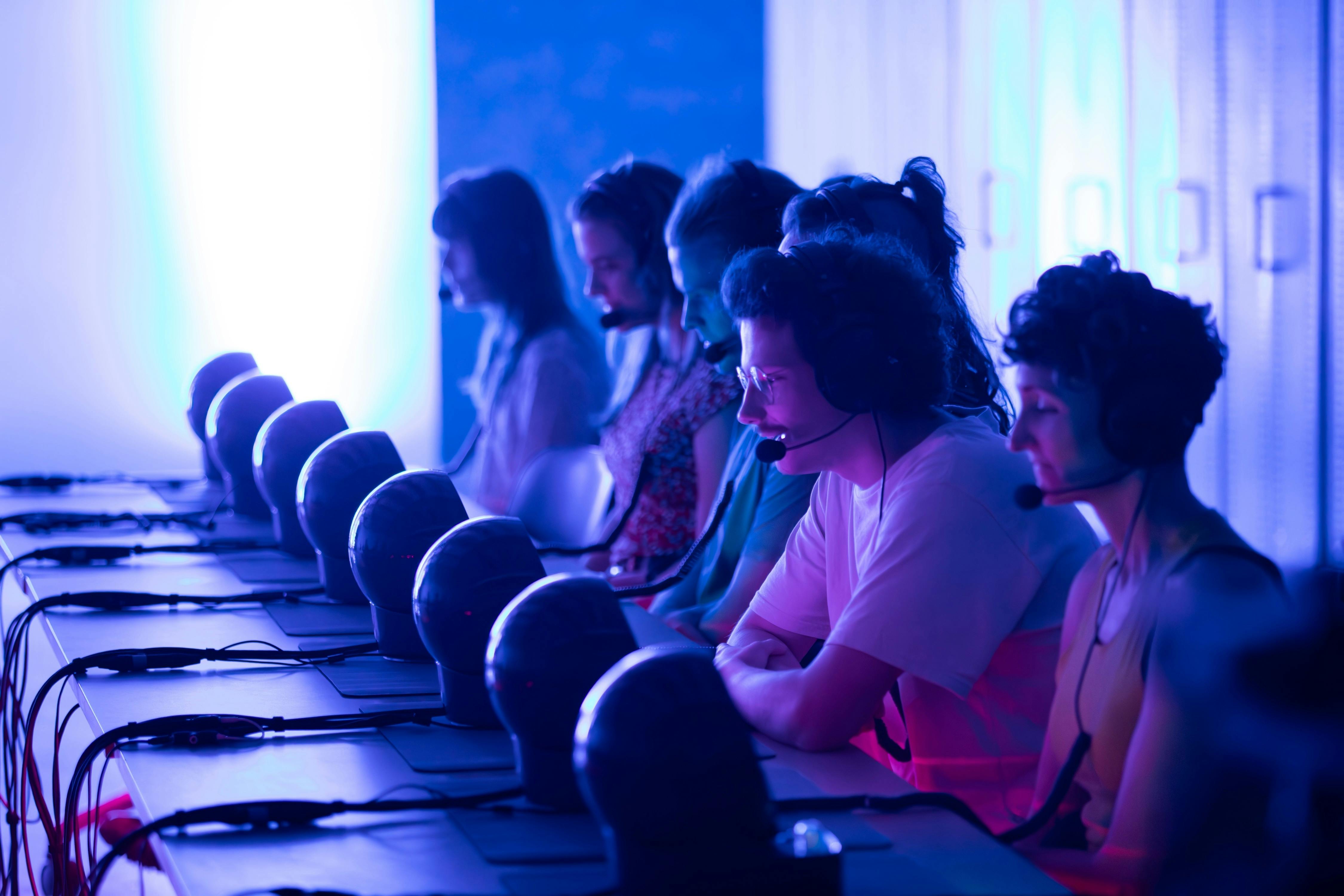 A row of people wearing headsets sit at computers in a dimly lit room with blue and pink lighting.