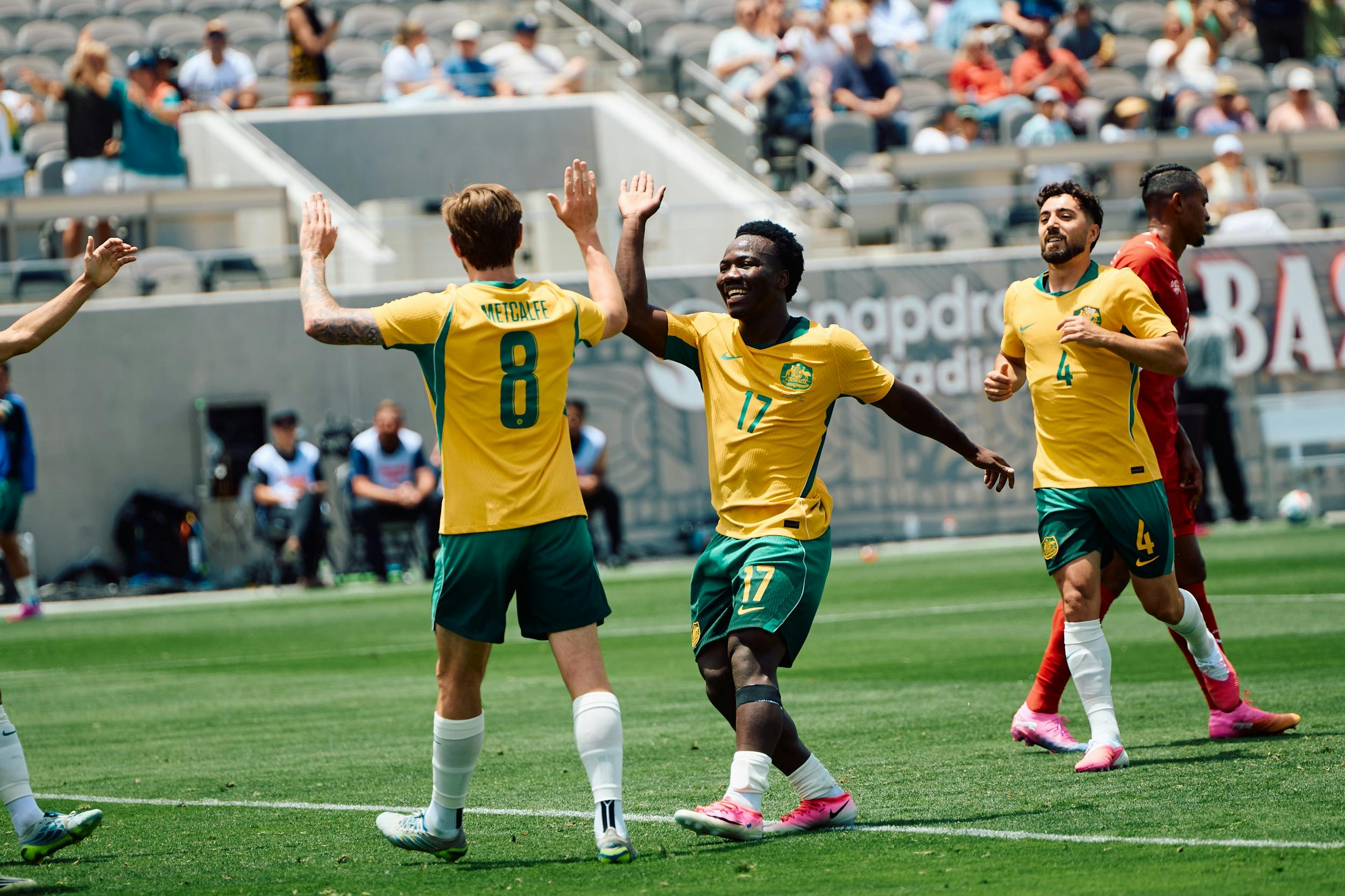 Soccer players in yellow kits celebrate a goal on a stadium pitch as teammates run in behind