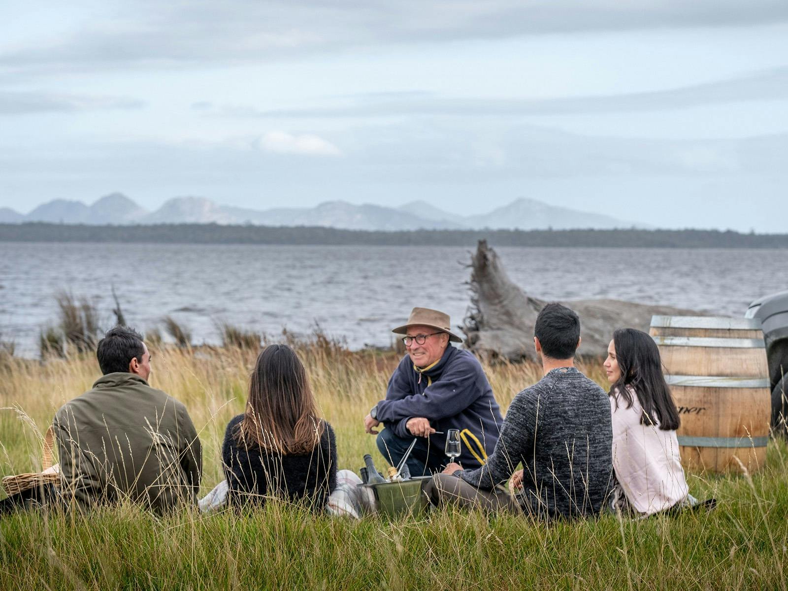 Group of people sitting in the grass overlooking water. Wine barrel is to their right.