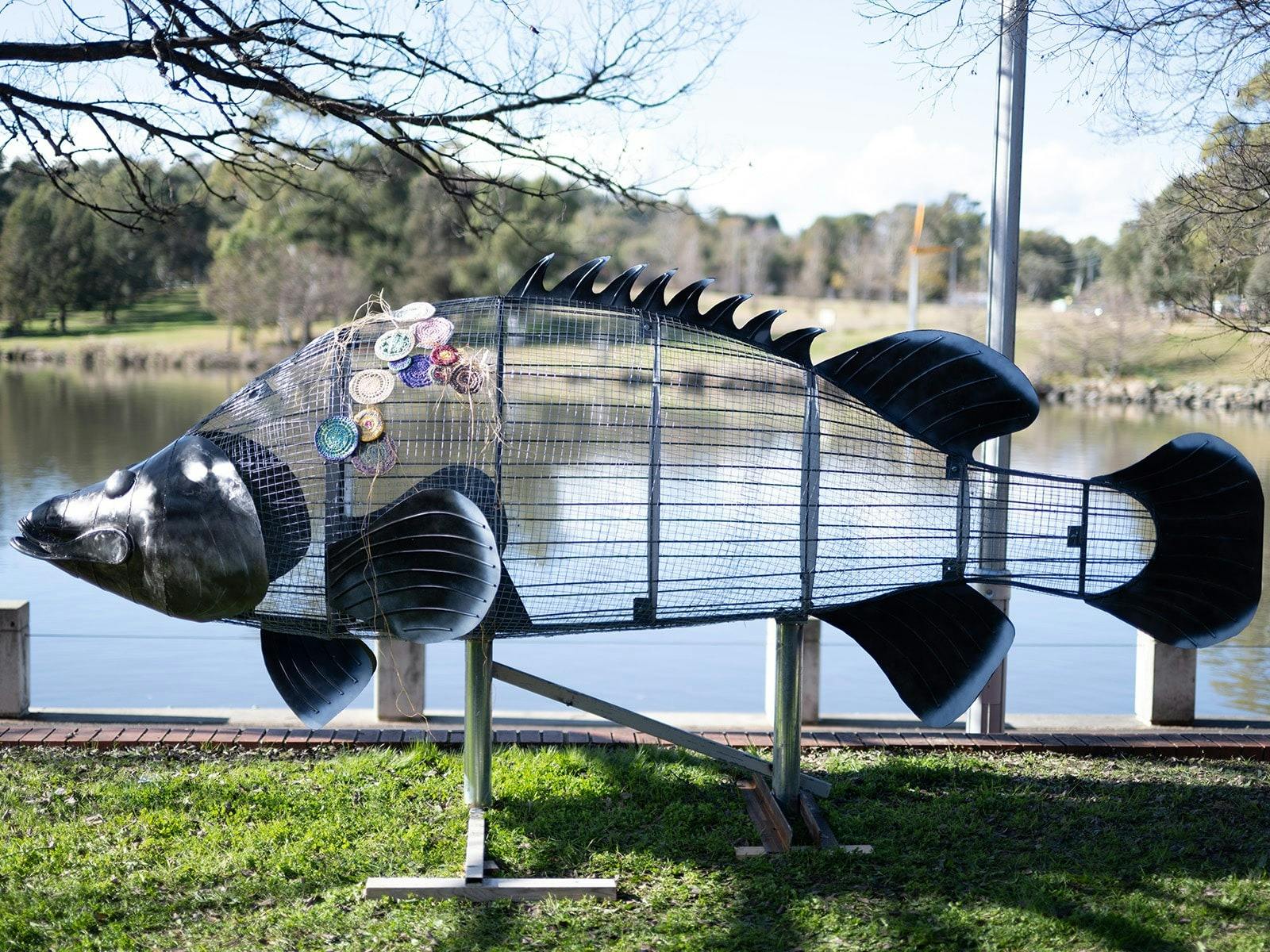 A steel wrieframe Cod sculpture with some woven textiles attached stands at the side of a lake