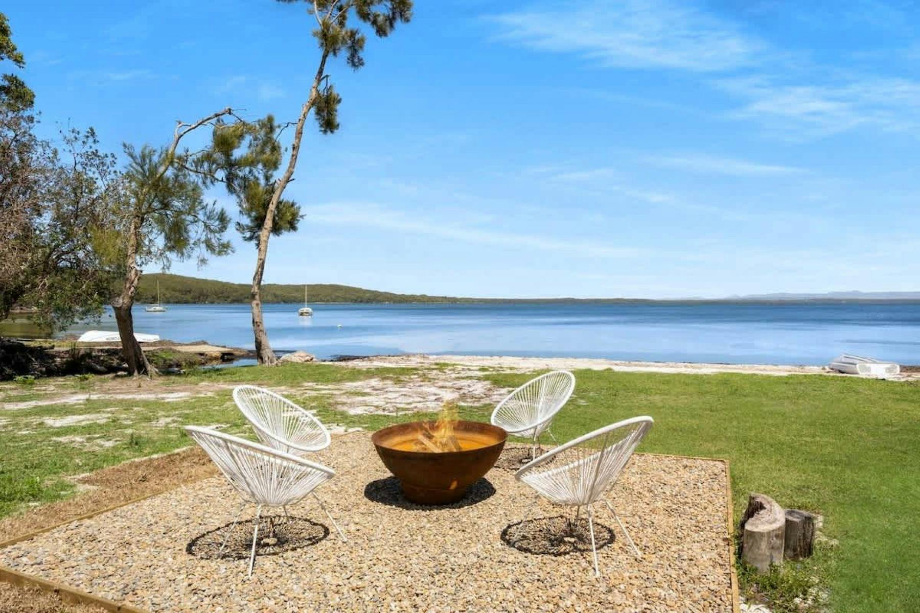 Firepit with backdrop of waterway and blue sky, tree sits to the left of the image. Fire is lit.