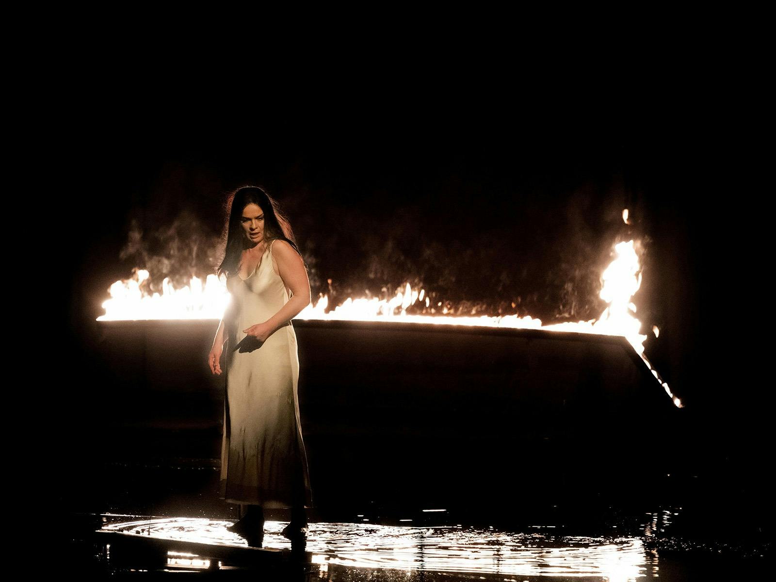 An opera singer on a dark stage wearing white, there is water pooled at her feet, fire behind her.