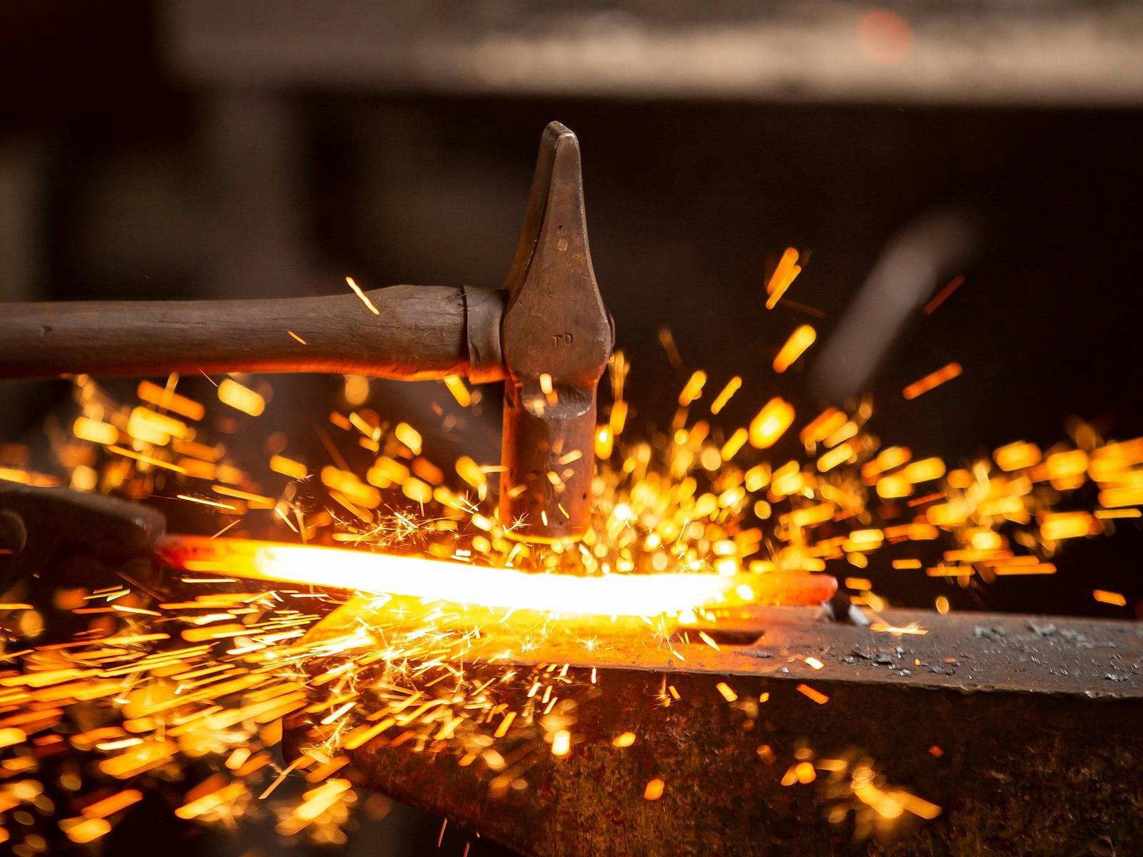 Hammer striking heated metal on anvil creating sparks.