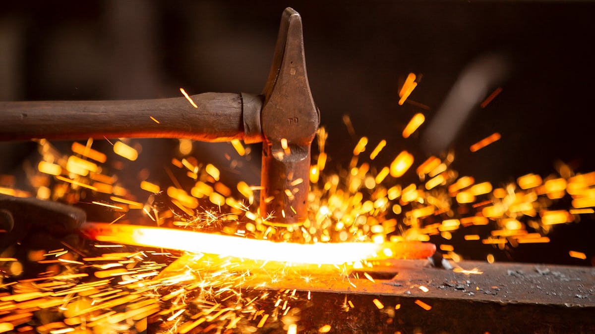Hammer striking heated metal on anvil creating sparks.