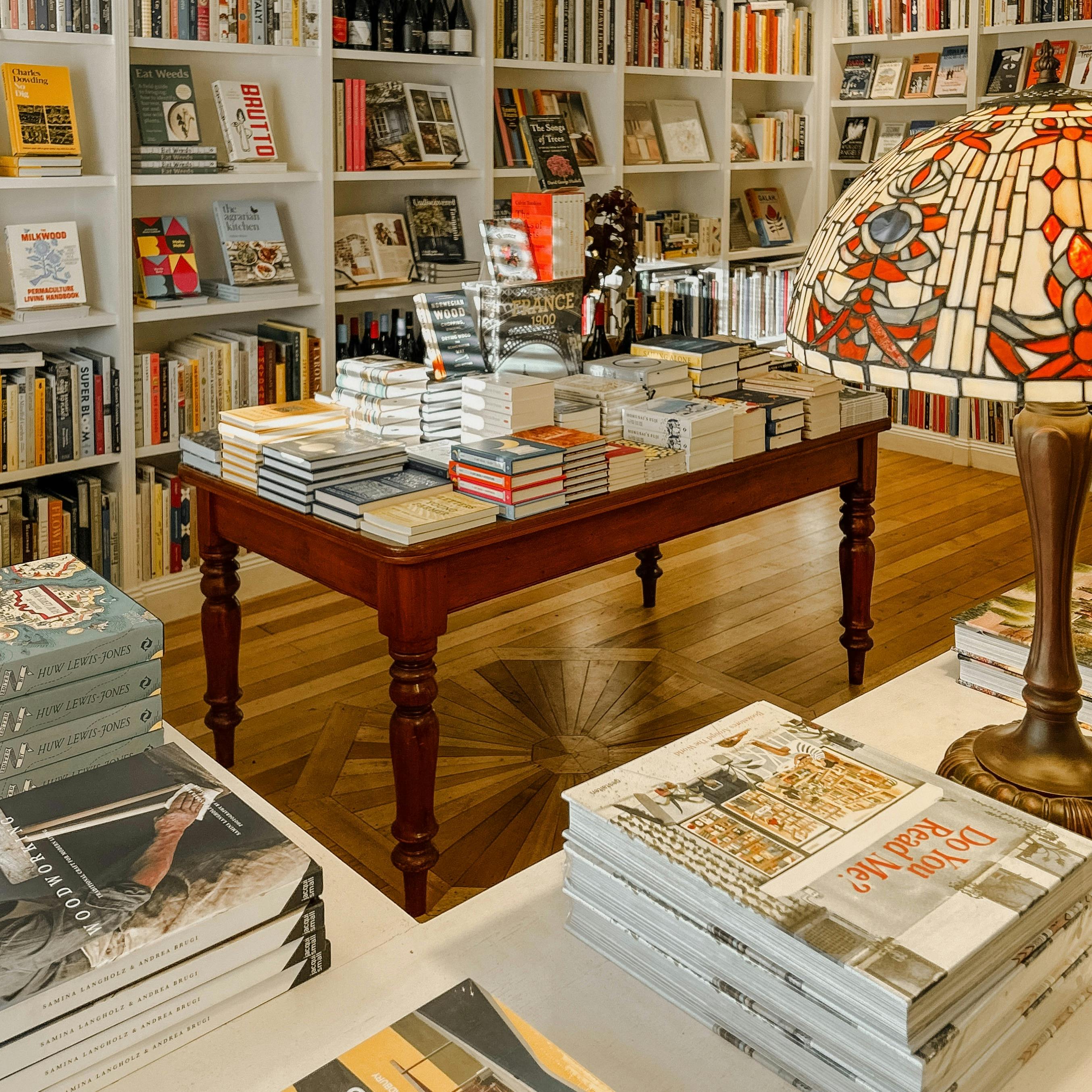 Table of stacked books in a cosy shop with shelves and a stained glass lamp nearby.