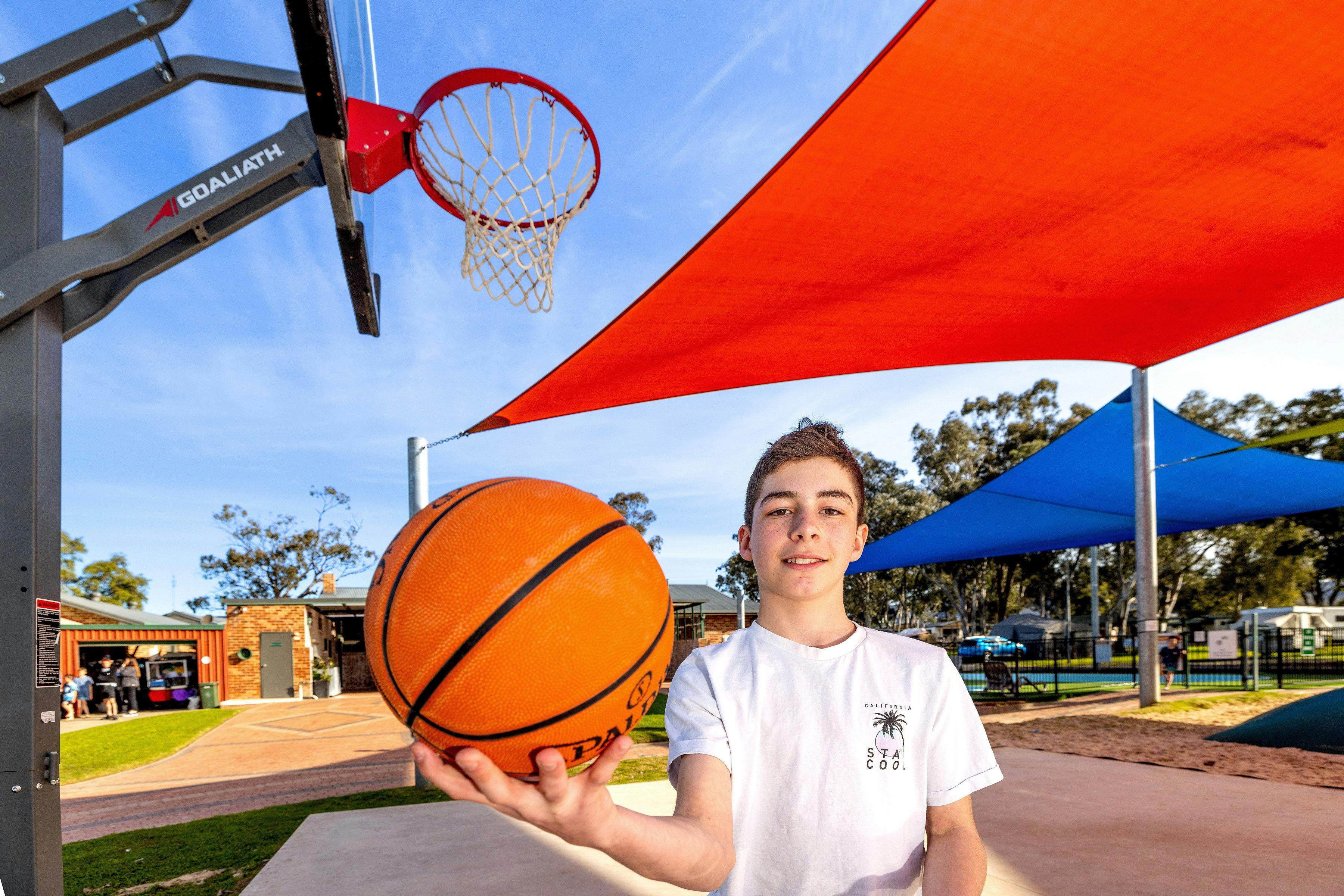 Boy holding a basket ball, with a basket ball hoop in the background.