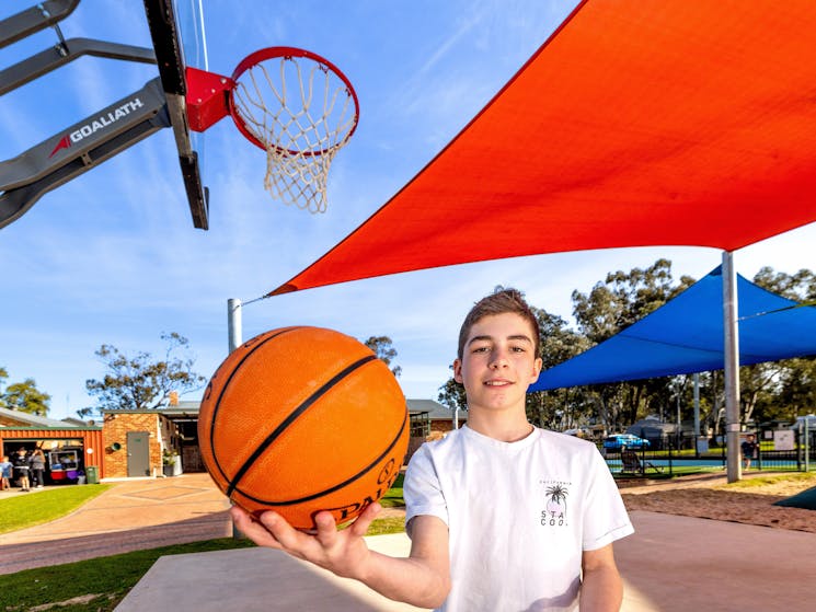 Boy holding a basket ball, with a basket ball hoop in the background.