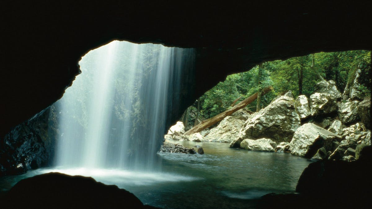 A sheet of white water, backlit by sunlight, falls into a dark rockpool,  underneath a rock arch.