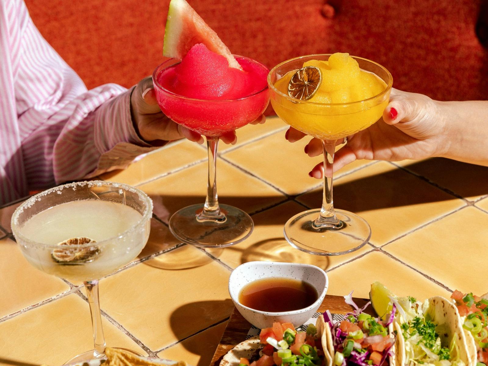 People cheersing a watermellon drink and orange drink with tacos down the bottom of image