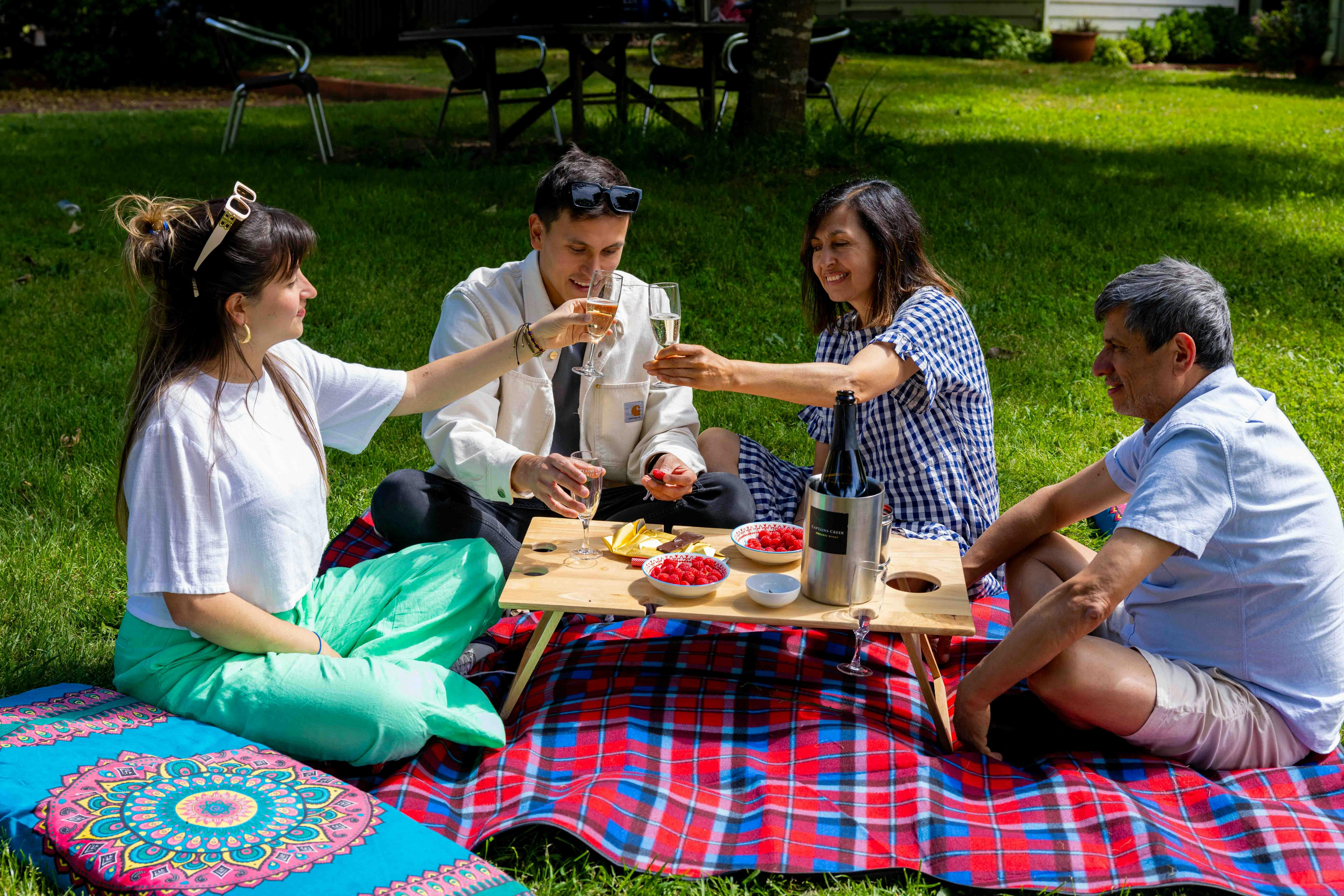 4 people sitting on tartan picnic rug with large cushions.  Dessert and sparkling wine on the table